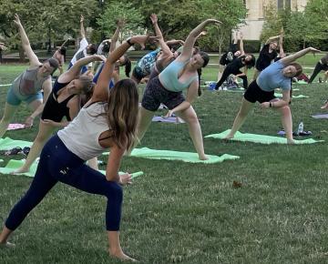 A group of young women do a yoga leaning pose outdoors on a grass lawn.
