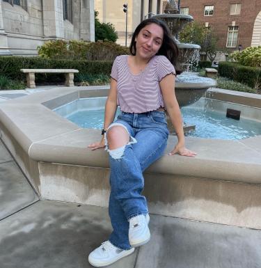 Photo of a smiling young woman in jeans with a knee tear and a short-sleeved stripy top sitting at a fountain.