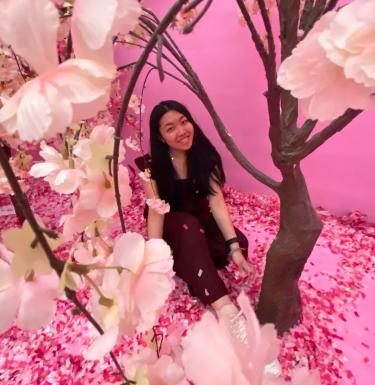 Photo of a smiling young woman sitting under a pink blossoming cherry tree art installation.