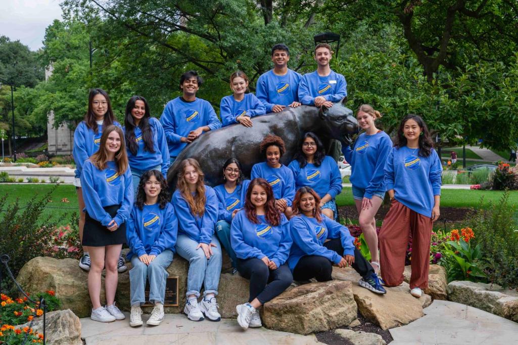 Outdoor group photo of about a dozen smiling students in matching blue Global Ties crewneck shirts around a panther statue.