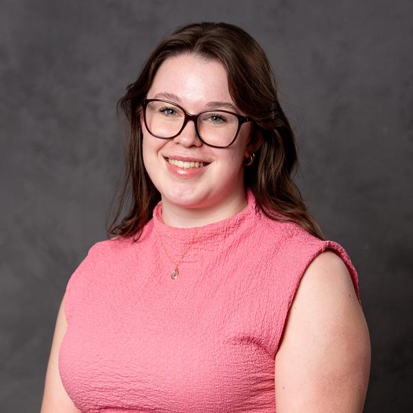 A woman with long dark hair and glasses smiling in a pink sleeveless top.