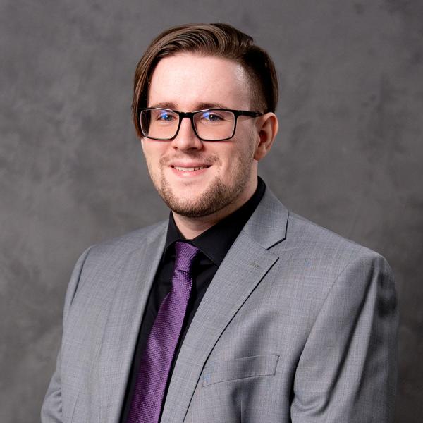 A man with short dark hair and glasses smiling in a light grey blazer over a purple button up shirt.