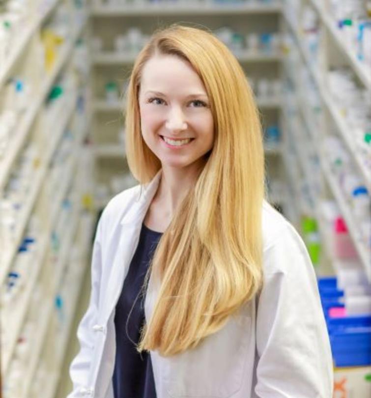 A smiling blonde woman in a white pharmacists's coat.