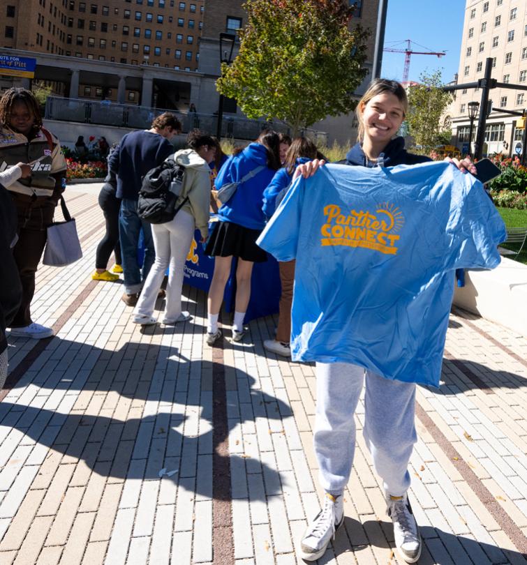 Student smiling holding a pale blue t-shirt up outdoors.