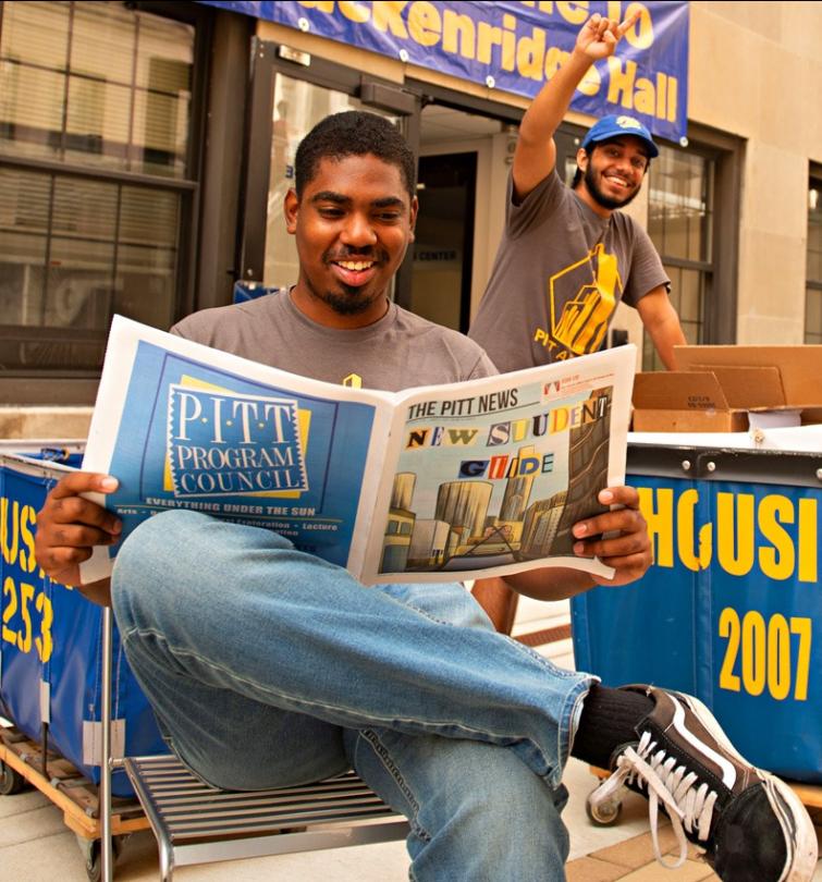 A smiling student sits and reads the Pitt newspaper outdoors as another waves at the camera and pushes a housing cart.