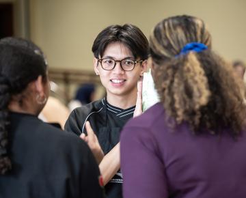 A male student with glasses speaking with two female students.
