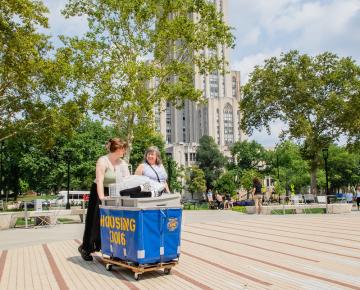 A mother and daughter pushing a blue housing cart past the Cathedral of Learning.