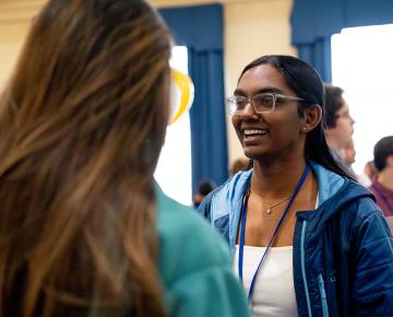 A female student in a blue zip up hoodie speaking to a student in a green top.