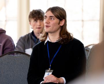 A male student with long brown hair sitting in a chair wearing a black sweater.