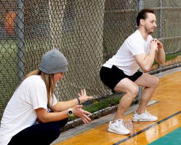 A male student performing squats while a staff member corrects their form.