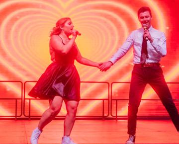 Students singing and holding hands onstage with a heart motif projected behind them.