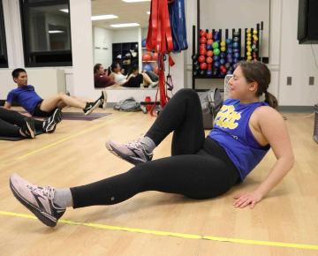 Smiling student in a blue Pitt tank top doing exercises on the floor of a gym.