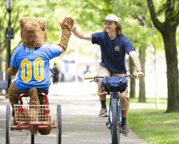 The Pitt Roc panther mascot on a bike high fives another bike rider outdoors.