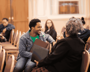 Smiling student with a folder talks with a staff member in a conference room with chairs arranged in rows.