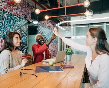 Three women in business causal garb high five over a wooden table.