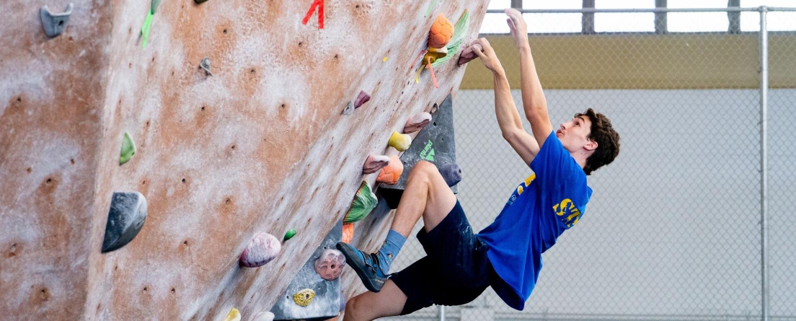 A male student in a blue Pitt tee shirt climbs a rock climbing wall without a harness.