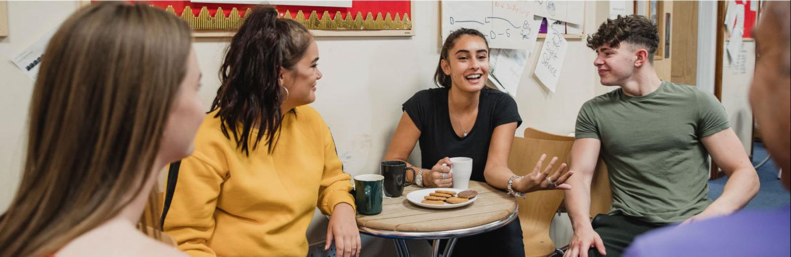 A group of four young students smile and talk in a classroom over drinks and cookies.