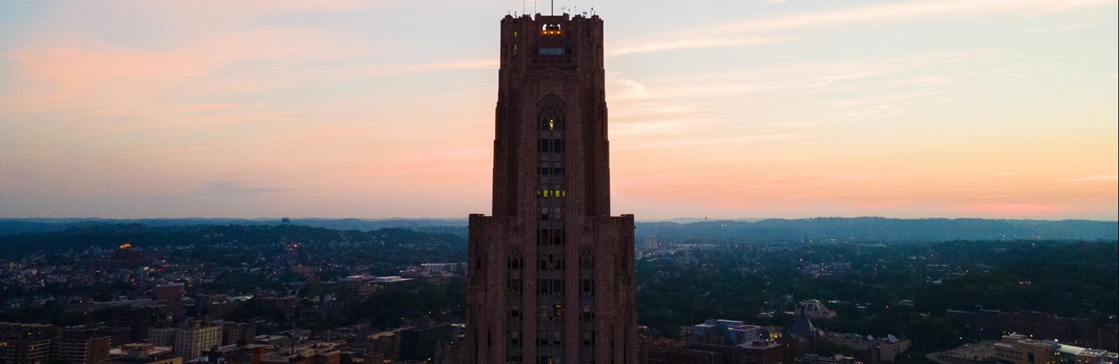 The Cathedral of Learning, a stately tan building rising over a city in front of a sunset.