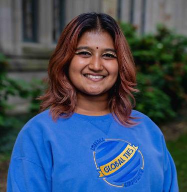 Headshot of a smiling person with rose gold dyed hair, gold eyeliner, a bindi, and wearing a blue Global Ties crewneck.