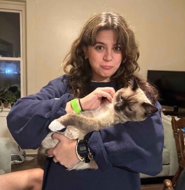 A young woman with brown curly hair and a playful expression holds and scratches a cream-colored Siamese cat.