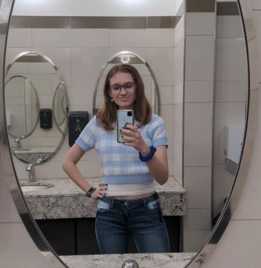 Mirror selfie of a young woman in a blue and white checkered short sleeve top with medium-length brown hair.