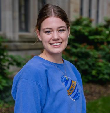 Outdoor headshot of a smiling woman in a blue Global Ties crewneck shirt and pearl earrings.