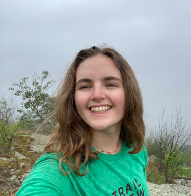 Outdoor selfie of a smiling young woman with wavy brown hair in a green tee shirt.