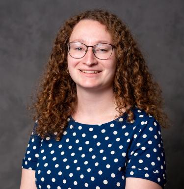 A woman with long curly hair and glasses smiling in a blue top with white polka dots.