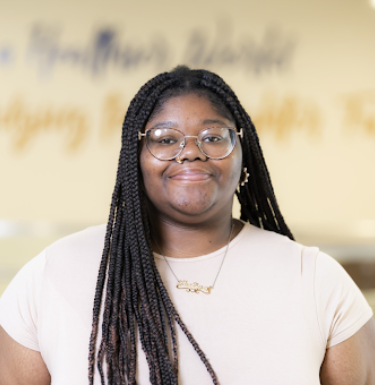 Headshot of a smiling woman in a black tee shirt, gold name necklace, and round glasses.