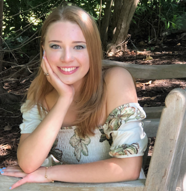 Headshot of a smiling young woman with strawberry-blonde hair sitting outside on a bench.