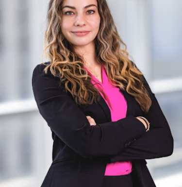 Business headshot of a young woman in a black blazer and hot pink shirt with curly blonde hair, arms crossed.