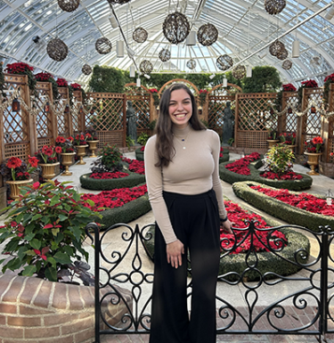 A woman smiling in a beige top and black pants in front of red flowers.