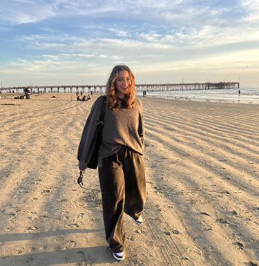 A woman with blonde hair in a grey sweater and black pants on a beach at sunset.