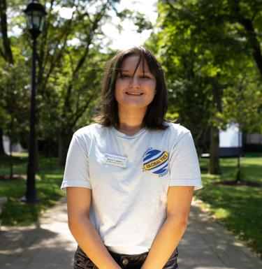 Headshot of a smiling woman with shoulder-length brown hair in a white Global Ties tee shirt standing outside.