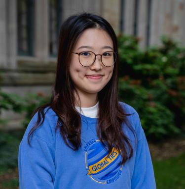 Headshot of a smiling woman in dark round glasses and a blue Global Ties crewneck shirt.