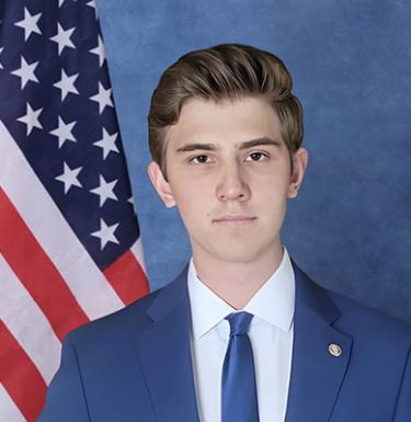 A man with short blonde hair in a blue suit standing in front of an American flag.