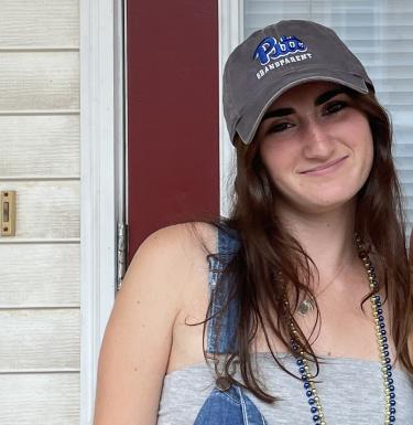 Photo of a young woman with long dark hair in a Pitt Grandparent cap, denim overalls, and blue and gold beads.