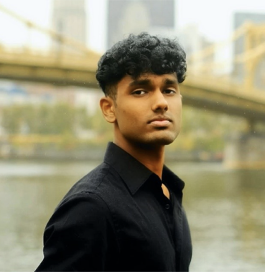 A man with short dark curly hair in a black shirt standing in front of a bridge.