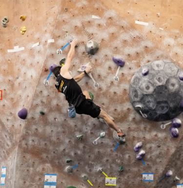 A man in a black tank top and black shorts climbing on a climbing wall.