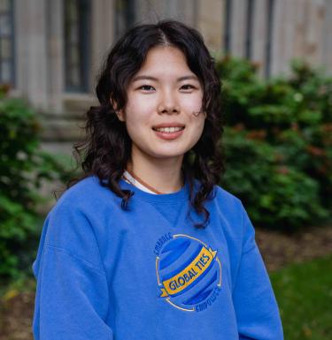 Headshot of a woman with curly dark brown hair standing outdoors in a blue Global Ties crewneck shirt.