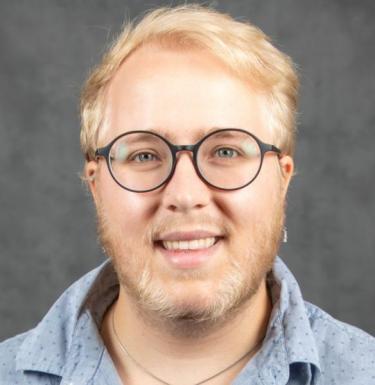 A person with short blonde hair and facial hair smiling with glasses on.