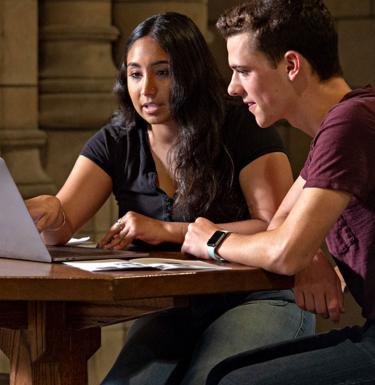A young man and woman look at a laptop on a wooden table.