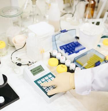 A gloved hand punches numbers into a small white device in a lab filled with bottles and tubes.