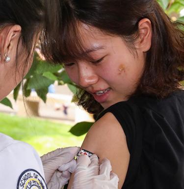 A woman in a white coat places a band-aid on another woman's arm outside.