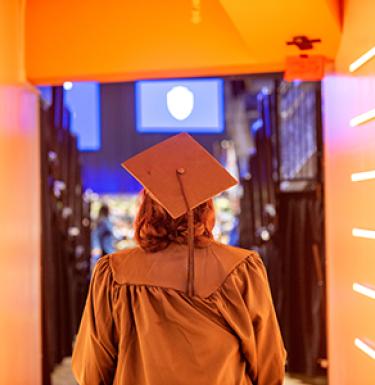 A student in a graduation cap and gown walking through a tunnel lit with neon lights.