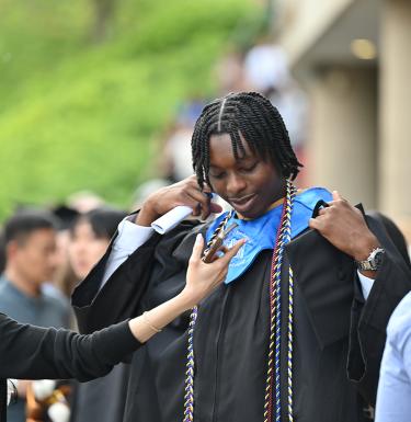 A Black student in commencement regalia receiving help with his hood.