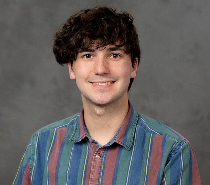 Brunette man smiling in a purple and green striped short sleeve button down shirt.