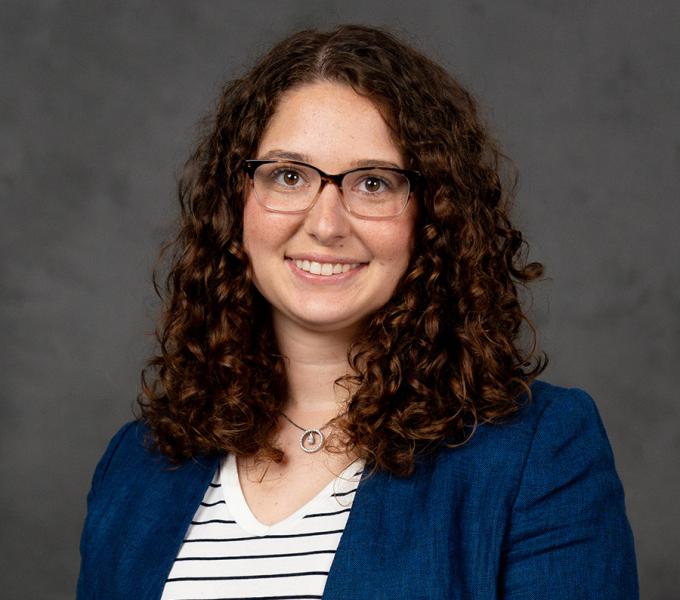 Woman with long dark curly hair with glasses smiling in a white and blue striped shirt under a navy blazer.