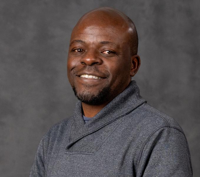 A bald Black man in a grey sweater smiling in front of a grey backdrop.