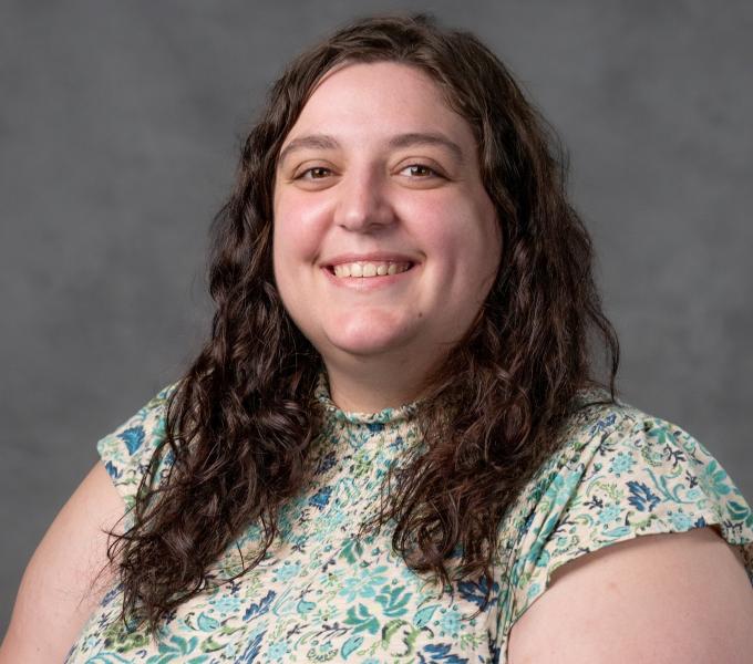 Headshot of a smiling woman with brown curly hair in a short-sleeved paisley shirt.